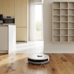 Entretien de la maison : ce qu'on peut automatiser et ce qu'il vaut mieux déléguer Intérieur familial lumineux avec un robot aspirateur circulant sur un parquet en bois, une cuisine bien rangée et un organisateur mural discret.