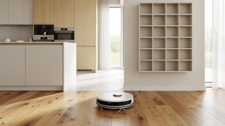 Intérieur familial lumineux avec un robot aspirateur circulant sur un parquet en bois, une cuisine bien rangée et un organisateur mural discret.